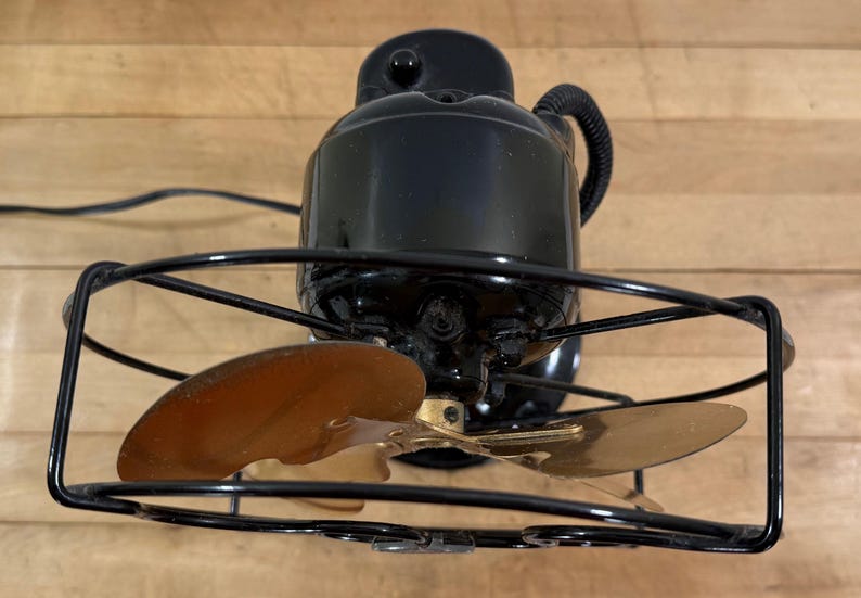 May include: Black electric fan with two bronze-coloured blades, encased in a black wire cage. The fan's motor housing is black, and the power cord is visible. The fan is set against a wooden background.