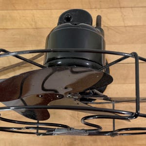 Pu&ograve; includere: Ventilatore elettrico nero vintage con tre pale e una gabbia protettiva. Il ventilatore ha un alloggiamento motore e pale neri e una gabbia in metallo nero. Il ventilatore &egrave; appoggiato su una superficie di legno.