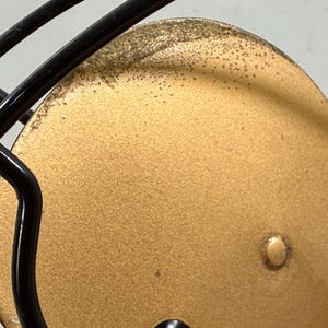May include: Close-up of a football helmet. The helmet has a gold-coloured face mask and a black metal cage. The gold surface has a textured appearance with small, dark spots.