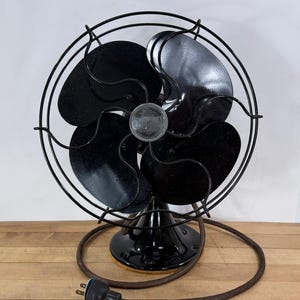Pu&ograve; includere: Ventilatore elettrico Emerson vintage con quattro pale nere e una gabbia in metallo nero. Il ventilatore ha una base nera e un cavo di alimentazione marrone con una spina a due poli. Il ventilatore &egrave; appoggiato su una superficie di legno.