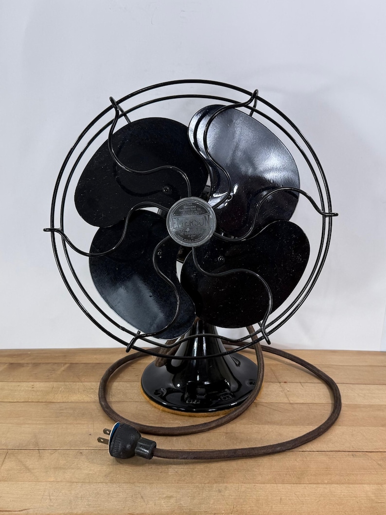 Pu&ograve; includere: Ventilatore elettrico Emerson vintage con quattro pale nere e una gabbia in metallo nero. Il ventilatore ha una base nera e un cavo di alimentazione marrone con una spina a due poli. Il ventilatore &egrave; appoggiato su una superficie di legno.