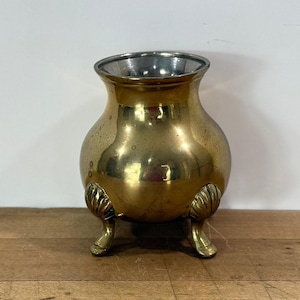 May include: A polished brass vessel with a rounded body and a wide, open top. The container features three distinct feet, each designed with a decorative shell-like motif at the top where it connects to the base of the rounded brass bowl.