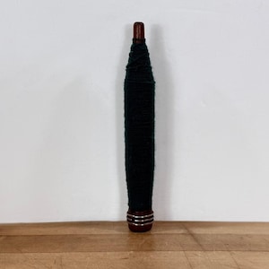May include: A dark green yarn swift, a tool used for winding yarn into a ball. The swift has a dark wooden top and bottom with silver metal rings. The yarn is tightly wound around the central wooden core.