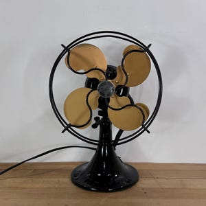 May include: An antique Emerson electric fan with four tan blades and a black metal frame. The fan has a black base and a circular cage. The fan is sitting on a wooden surface.