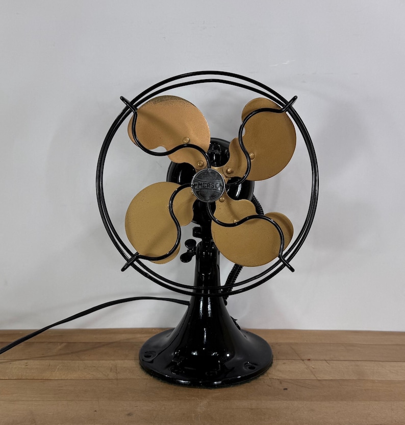 May include: An antique Emerson electric fan with four tan blades and a black metal frame. The fan has a black base and a circular cage. The fan is sitting on a wooden surface.