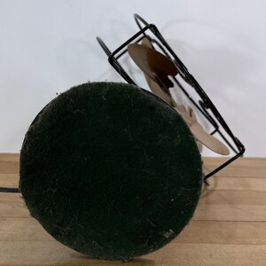 May include: A vintage desk fan featuring a black metal frame and three tan blades. The fan has a round, dark green felt base and a black power cord. The fan is angled upwards, displaying its design.