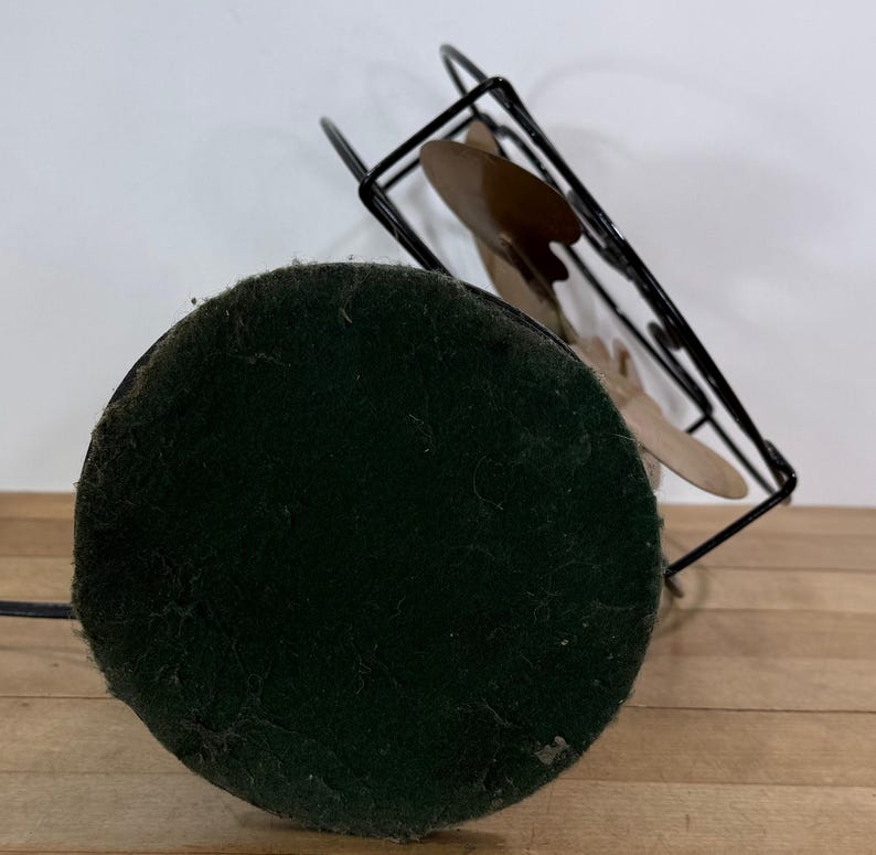May include: A vintage desk fan featuring a black metal frame and three tan blades. The fan has a round, dark green felt base and a black power cord. The fan is angled upwards, displaying its design.
