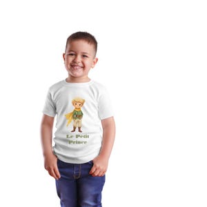 Puede incluir: Una camiseta blanca con una ilustraci&oacute;n de dibujos animados del Principito, que lleva una bufanda amarilla y sostiene una caja. El texto "Le Petit Prince" est&aacute; impreso debajo de la ilustraci&oacute;n.