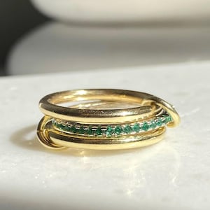 May include: Three gold rings stacked together. The middle ring features a line of small, green gemstones. The rings are presented on a white surface, with a blurred background.