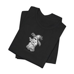 May include: Black t-shirt featuring a grayscale illustration of a ghost wearing a sombrero and playing a guitar. The ghost has a halo and is dressed in a poncho. The shirt is folded and has a simple, clean design.