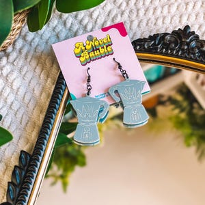 May include: A pair of light blue coffee pot-shaped earrings with black hooks. The earrings are displayed on a pink tag with the text "A Novel Bauble" in a retro font. The earrings are reflected in a mirror.