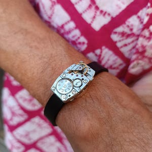 Op de afbeelding: Een zilveren horloge met een zwarte band. De wijzerplaat van het horloge is blootgesteld en laat het innerlijk van het horloge zien.