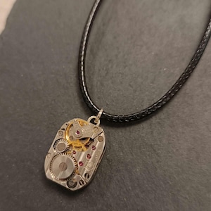 Op de afbeelding: Een zilverkleurige hangerketting met de ingewikkelde binnenkant van een horloge. De rechthoekige hanger toont tandwielen, juwelen en andere mechanische componenten. De ketting heeft een zwart leren koord.