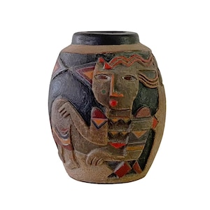 May include: A ceramic vase with a dark brown rim and a textured, earthy body. The vase features a carved design of a stylized human figure with geometric patterns in red, orange, and black. The design is reminiscent of tribal art. The vase is approximately 20 cm tall.