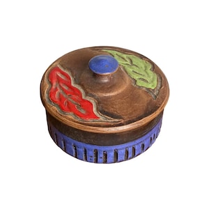 May include: A round ceramic lidded jar with a brown base and a blue lower section featuring vertical indentations. The lid has a central blue knob, a red leaf-shaped relief, and a green leaf-shaped relief on a brown textured surface.