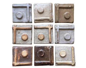 May include: A collection of nine square ceramic tiles arranged in a three-by-three grid. Each tile features a raised circular center and a raised L-shaped border along two edges. The tiles display various earth-toned glazes including gray, tan, and brown.