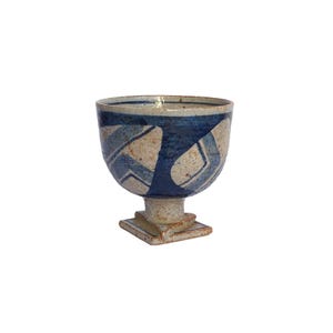 Pode incluir: Um cálice de cerâmica com uma taça larga e arredondada e uma base quadrada. O cálice apresenta um esquema de cores bege e azul salpicado com padrões geométricos azuis. A borda e o interior são de um azul mais escuro.