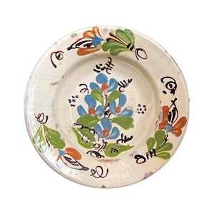 Könnte beinhalten: Ein weißer Keramikteller mit handbemaltem Blumendesign. Die Blumen sind blau, grün und orange. Der Teller hat einen rustikalen, Vintage-Look.