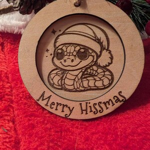 Może przedstawiać: Okrągła drewniana ozdoba choinkowa z laserowo grawerowanym obrazkiem węża w czapce Świętego Mikołaja. Ozdoba ma napis "Merry Hisssmas".