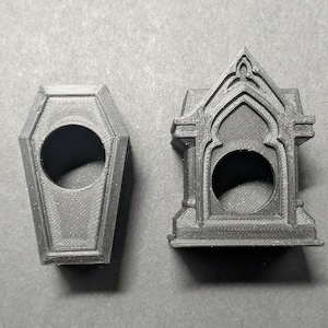 Op de afbeelding: Twee zwarte 3D-geprinte items: een object in de vorm van een kist en een miniatuur gotische boog. Beide hebben een cirkelvormige opening. De kist is rechthoekig met een afgeronde bovenkant.