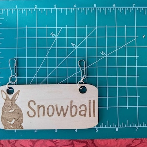 May include: A wooden sign with the name "Snowball" engraved on it. The sign has a brown rabbit illustration and two metal rings for hanging.