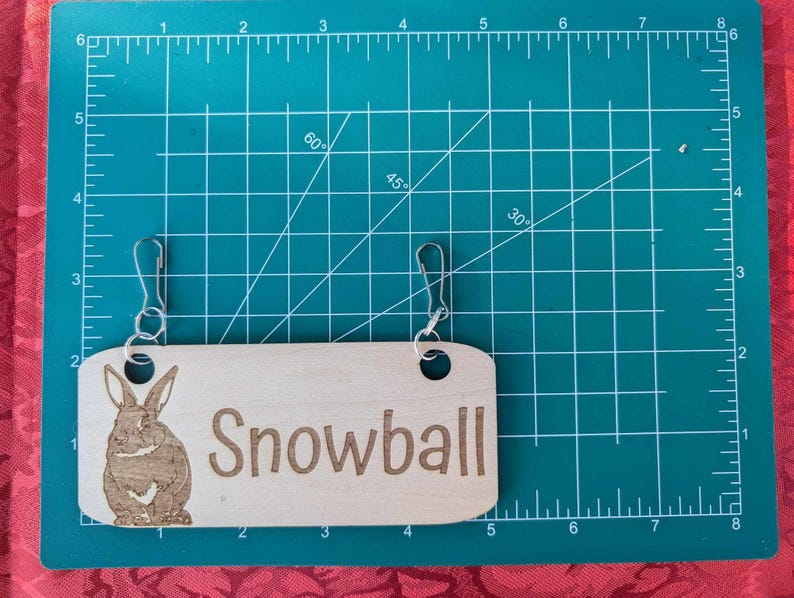 May include: A wooden sign with the name "Snowball" engraved on it. The sign has a brown rabbit illustration and two metal rings for hanging.