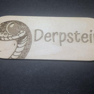 May include: A rectangular wooden plaque with rounded corners. The plaque features a detailed engraving of a snake's head on the left side and the name "Derpstein" in a playful font on the right. The wood has a light, natural color.