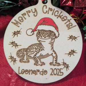 Peut inclure: Ornement de Noël rond en bois avec un gecko portant un bonnet de Père Noël, avec le texte "Merry Crickets!" et "Leonardo 2025". L'ornement a un trou pour l'accrochage et des détails gravés au laser.