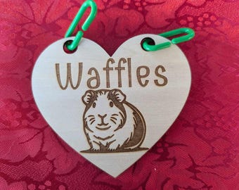 May include: A wooden heart-shaped tag with the name "Waffles" engraved on it. A small guinea pig is also engraved on the tag. The tag has two green plastic clips attached to it.