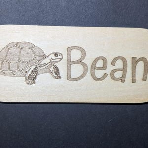 May include: A light-colored wooden sign with rounded corners. A detailed engraving of a turtle is on the left, and the name "Bean" is engraved on the right. The sign is set against a dark background.