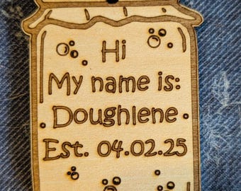 May include: A wooden jar-shaped tag with the text "Hi My name is: Doughlene. Est. 04.02.25" and small circles around the text.