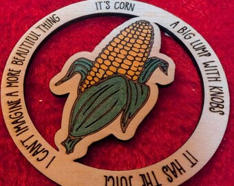 Puede incluir: Adorno de madera con un gráfico de maíz y el texto "IT'S CORN" y "I CAN'T IMAGINE A MORE BEAUTIFUL THING". El adorno también incluye las frases "A BIG LUMP WITH KNOBS" y "IT HAS THE JUICE".