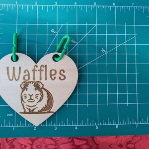 May include: A wooden heart-shaped tag with the word "Waffles" engraved on it. The tag is attached to a green string and features a laser-engraved image of a guinea pig.