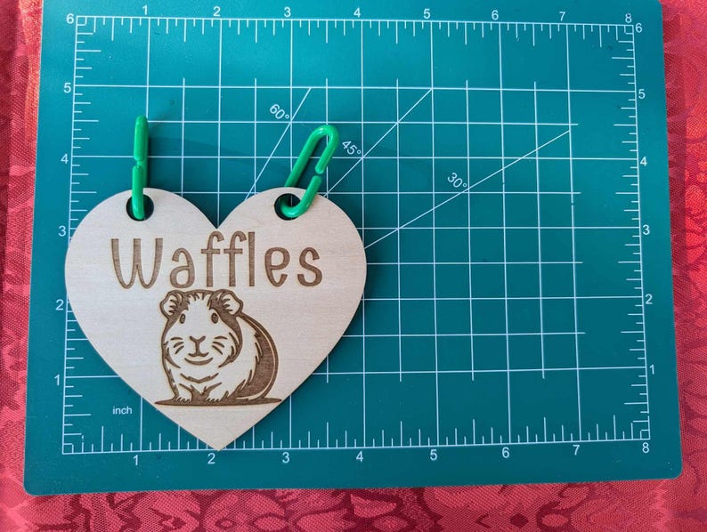 May include: A wooden heart-shaped tag with the word "Waffles" engraved on it. The tag is attached to a green string and features a laser-engraved image of a guinea pig.