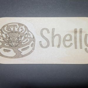 Op de afbeelding: Een lichtgekleurd houten bord met afgeronde hoeken. Het bord heeft een gedetailleerde gravure van een slang aan de linkerkant en de naam "Shelly" in een speels lettertype aan de rechterkant.