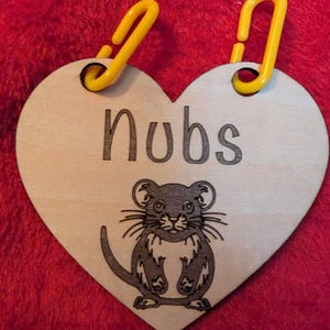 May include: A wooden heart-shaped tag with a black and white illustration of a mouse. The tag has two yellow plastic loops at the top and the word "Nubs" written in black.