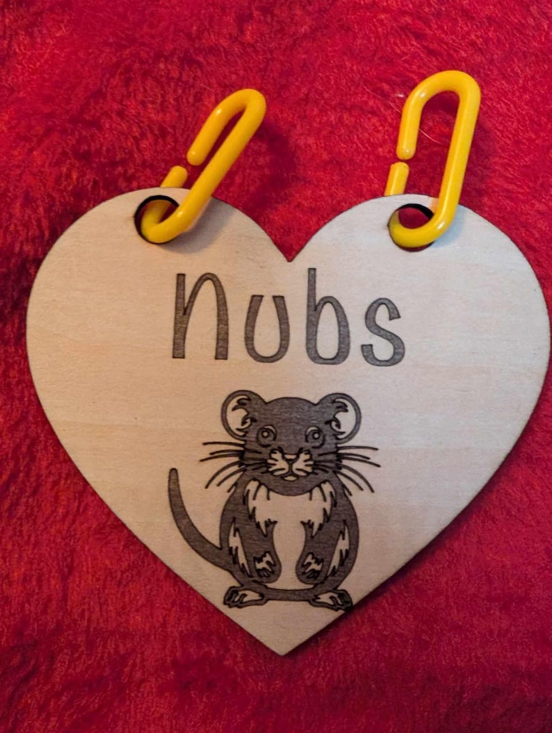 May include: A wooden heart-shaped tag with a black and white illustration of a mouse. The tag has two yellow plastic loops at the top and the word "Nubs" written in black.