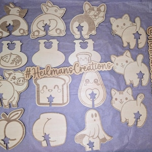 May include: A collection of wooden cutouts in various shapes, including animals, food items, and a ghost, each with a star-shaped cutout. The cutouts are light brown and arranged on a purple surface. The text "#HeilmansCreations" is visible.