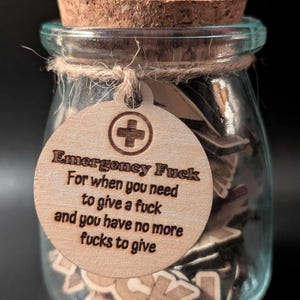Op de afbeelding: Een heldere glazen pot met een kurken stopper en een houten label. Het label leest "Emergency Fuck" en bevat de tekst "For when you need to give a fuck and you have no more fucks to give." De pot bevat kleine houten lettertegels.