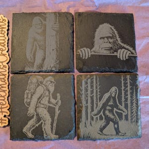 Könnte beinhalten: Set aus vier quadratischen Schieferuntersetzern mit geätzten Bigfoot-Designs. Jeder Untersetzer zeigt eine andere Bigfoot-Illustration, darunter eine, die über eine Kante schaut, und eine andere, die durch einen Wald geht. Die Untersetzer sind dunkelgrau.