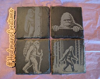 Peut inclure: Ensemble de quatre dessous de verre carrés en ardoise, chacun gravé d'un motif de Bigfoot différent. Les dessous de verre sont gris foncé et présentent diverses illustrations de Bigfoot, dont un regardant par-dessus un rebord.
