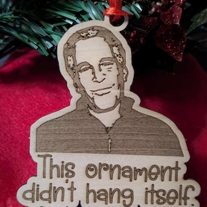 Może przedstawiać: Drewniana ozdoba choinkowa z portretem i napisem "This ornament didn't hang itself." Ozdoba jest przymocowana do czerwonej wstążki do zawieszenia. Tło zawiera dekoracje świąteczne.