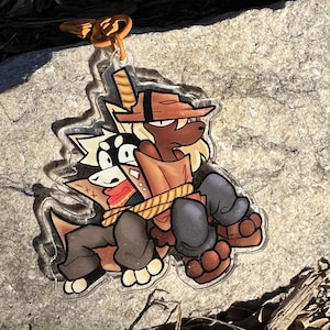 Op de afbeelding: Een heldere acryl sleutelhanger met een cartoon illustratie van twee antropomorfe dierenfiguren. Eén personage draagt een bruine hoed en jas, de andere heeft zwart-witte vacht. De sleutelhanger heeft een goudkleurige sluiting.