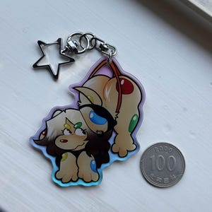 Op de afbeelding: Een heldere acryl sleutelhanger met twee cartoonfiguren en een stervormige bedel. De karakters zijn bruin, bruin en blauw met een holografische rand. Een 100-won munt ligt naast de sleutelhanger.