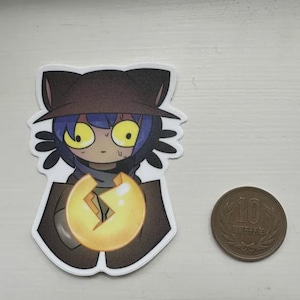 May include: A sticker featuring a cartoon character with blue hair, large yellow eyes, and a brown hat. The character is holding a glowing, cracked orb. A Japanese 10-yen coin is next to the sticker.