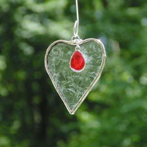 May include: A heart-shaped glass ornament with a silver-colored metal frame and a red teardrop-shaped gem. The glass has a textured, crackled appearance. The ornament hangs from a silver-colored wire against a blurred green background.
