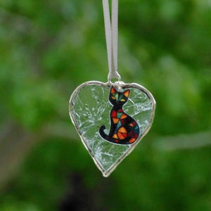 May include: A heart-shaped ornament featuring clear crackle glass encased in a silver metal frame. A small, colorful cat silhouette with a mosaic-style pattern of red, orange, yellow, and blue segments is attached to the center. It hangs from a white ribbon.