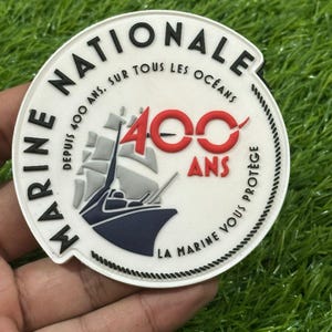 Könnte beinhalten: Ein kreisförmiger weißer Aufnäher mit dem Text MARINE NATIONALE, DEPUIS 400 ANS, SUR TOUS LES OCÉANS, 400 ANS und LA MARINE VOUS PROTÈGE. Das Design zeigt ein Segelschiff in Blau und Grau mit der roten Zahl 400 in der Mitte.