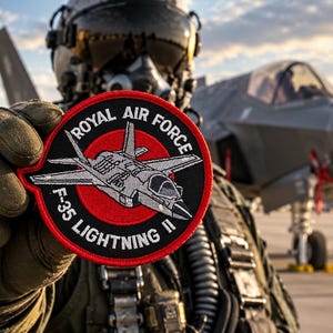 May include: A circular embroidered patch featuring a red center circle with a gray F-35 Lightning II aircraft illustration, surrounded by a black border with the white text ROYAL AIR FORCE and F-35 LIGHTNING II stitched in a bold, sans-serif font.