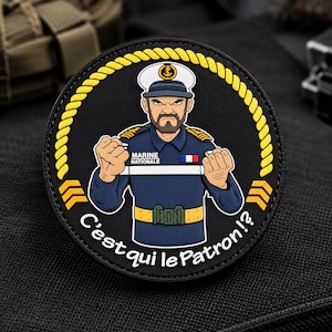 May include: A circular black patch featuring a cartoon illustration of a naval officer in a blue uniform with a white cap, gold epaulets, and a French flag pin. The text reads MARINE NATIONALE and C'est qui le Patron!? framed by a yellow rope border.
